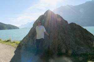 Robin Wils stands in front of a large rock by a lake with mountains in the background on a sunny day.